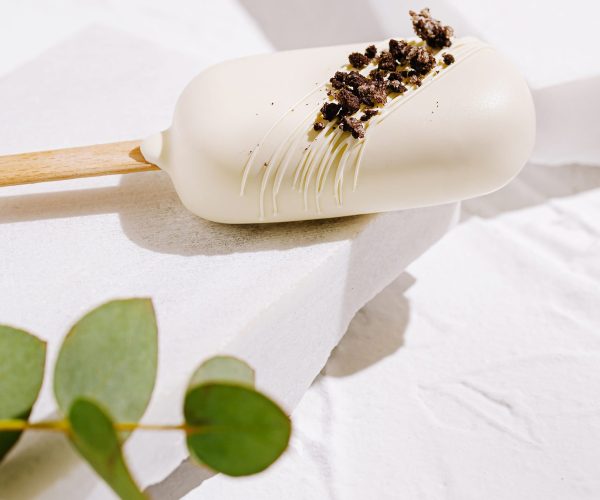 A creamy ice cream popsicle rests on a white surface. It features a crunchy topping and is accompanied by fresh green eucalyptus leaves, creating a refreshing aesthetic.