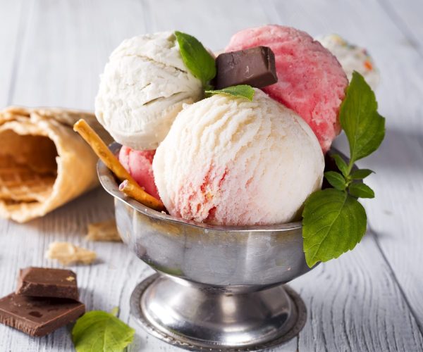vanilla and pink ice cream in a pial with frozen berries, chocolate and waffle cone on light wooden background